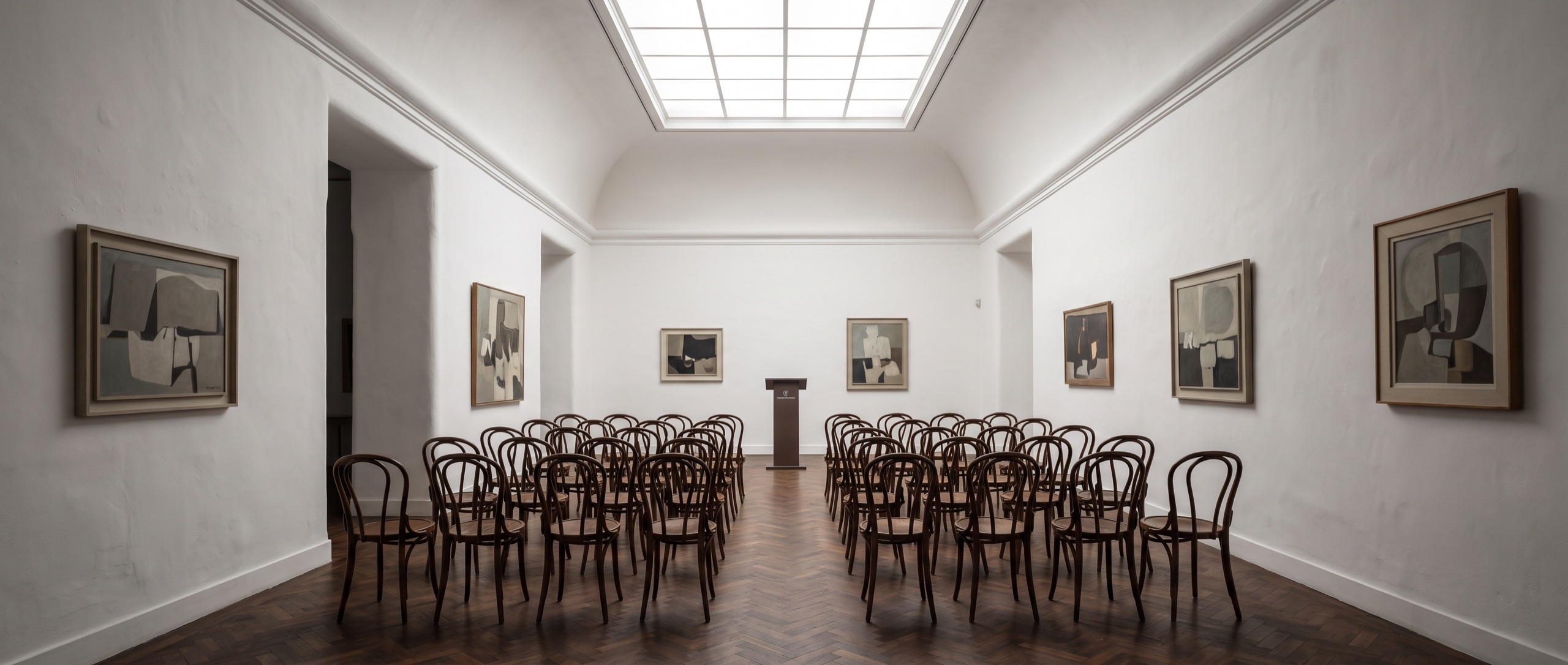 Sala de exposição da Casa Almeida: pinturas modernistas em paredes de gesso branco, soalho escuro, clarabóia com luz de norte e filas de cadeiras vazias.