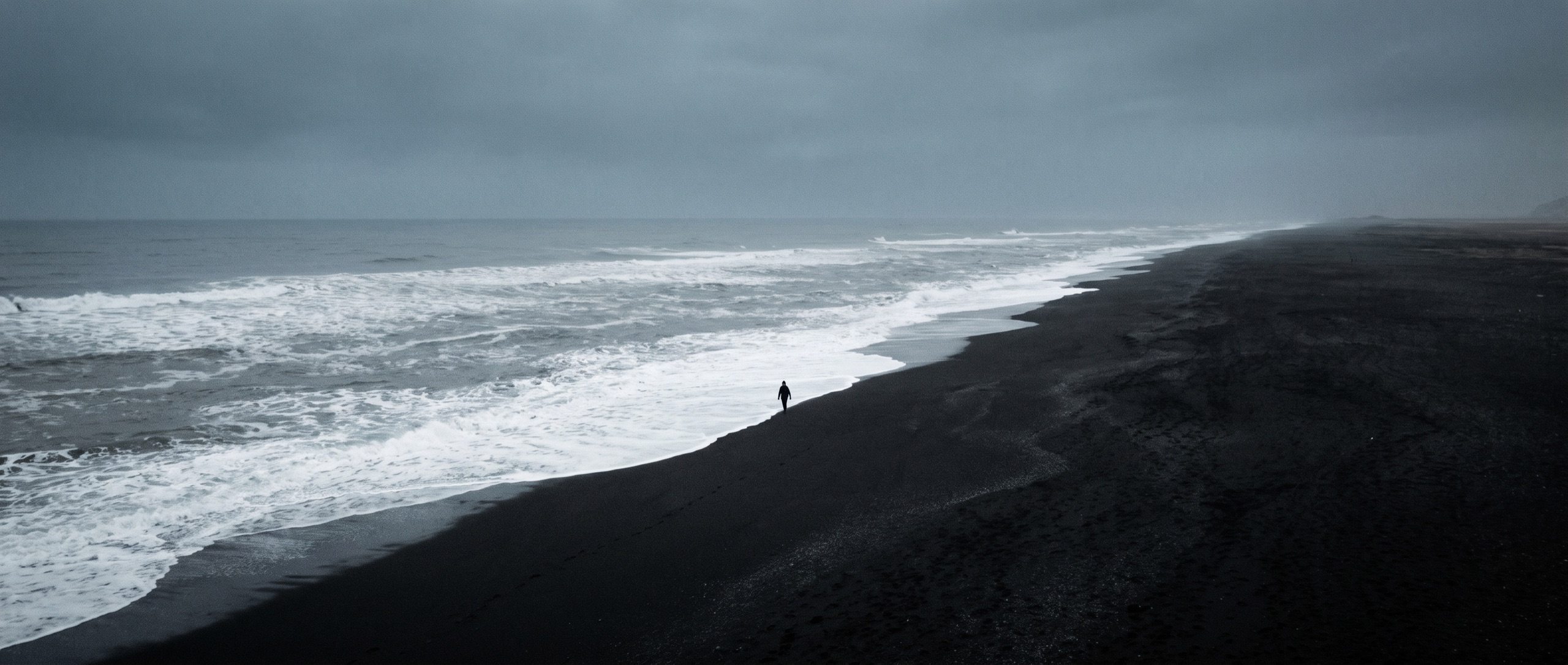 Aerial view of a black volcanic beach at dawn, a lone figure walking the shoreline