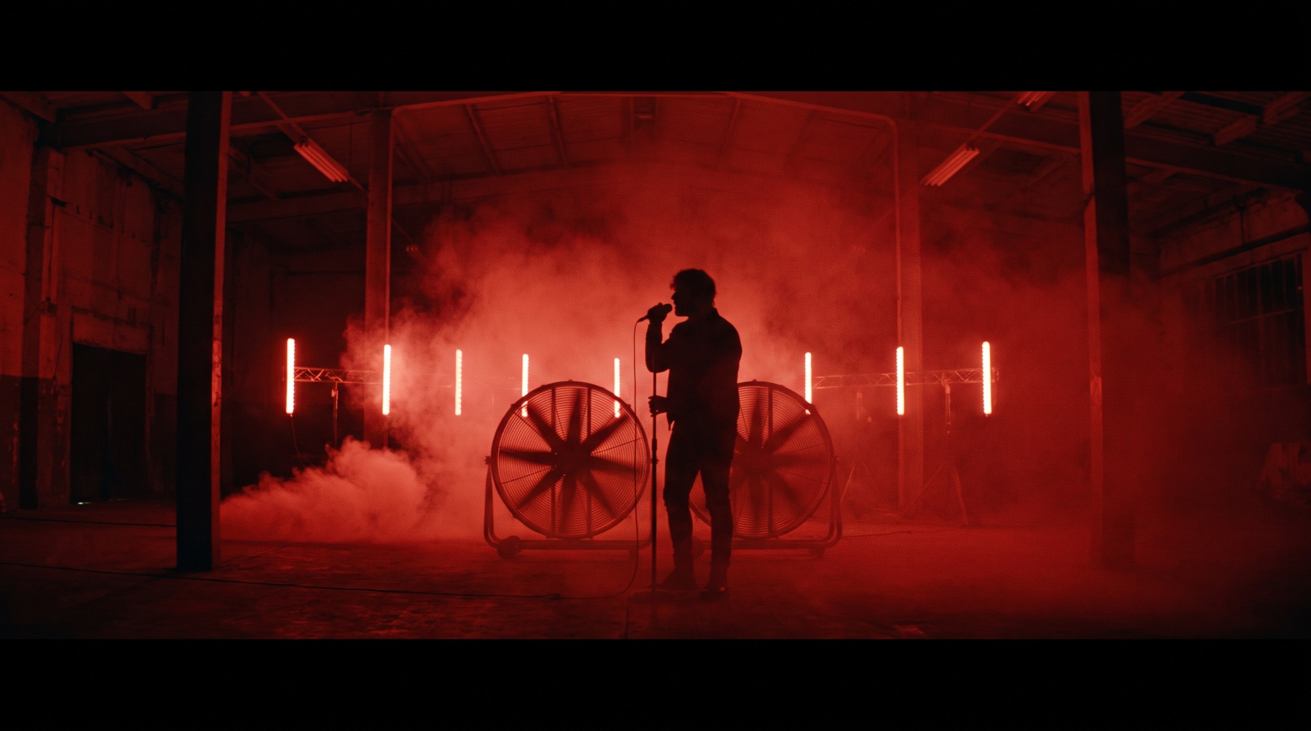 Singer silhouetted in red haze with industrial fans and tube lights